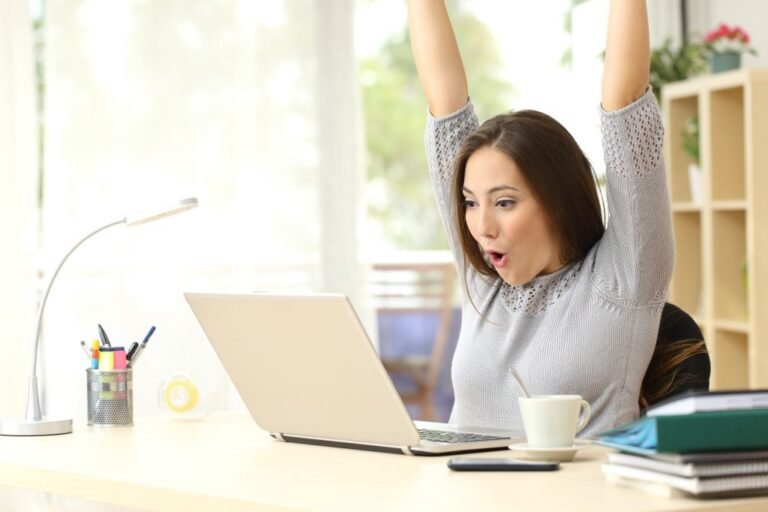 A woman is joyfully celebrating her online business success with her hands raised in front of a laptop. She is a digital marketing expert in Pakistan